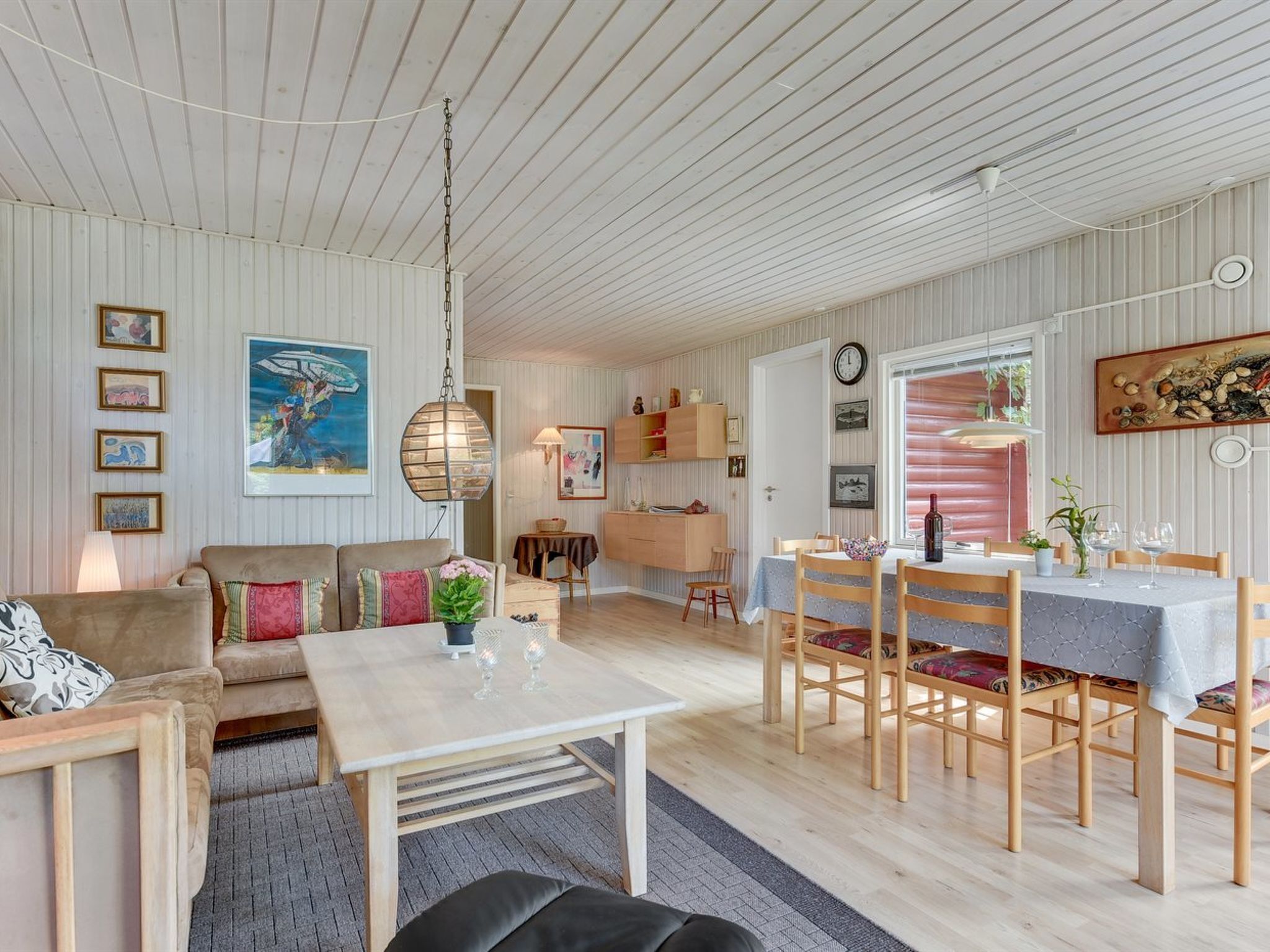 A cozy, well-lit room with a wooden ceiling, a dining table set for four, and a sofa with cushions.