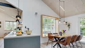 Une cuisine moderne dotée d’un îlot central, d’une table à manger en bois et de chaises. On y trouve des luminaires suspendus, une fenêtre offrant une vue agréable et un comptoir de cuisine sur lequel sont disposés divers objets.