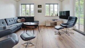 Un salon moderne comprenant un canapé d’angle, une table basse ronde, un fauteuil en cuir noir, un petit pouf et un téléviseur fixé au mur.