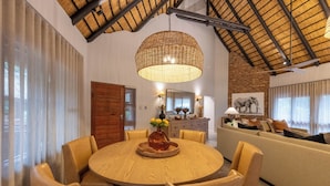 A dining area with a round wooden table and chairs, a hanging rattan light fixture, a brick wall, and a sofa with cushions.