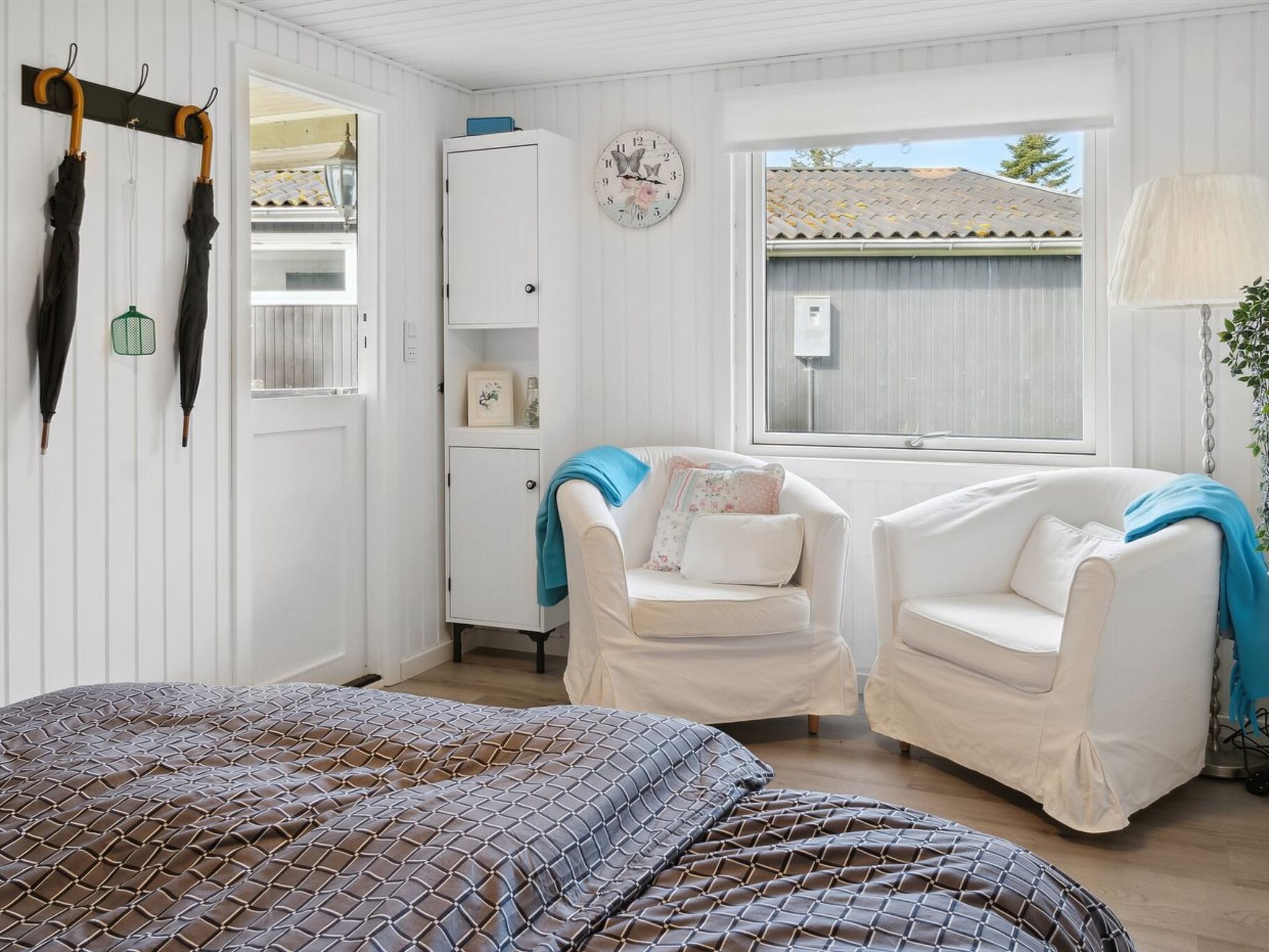 Une pièce confortable avec un lit, deux fauteuils blancs, une petite armoire et une fenêtre donnant sur un immeuble.