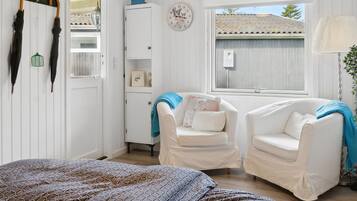 Une pièce confortable avec un lit, deux fauteuils blancs, une petite armoire et une fenêtre donnant sur un immeuble.