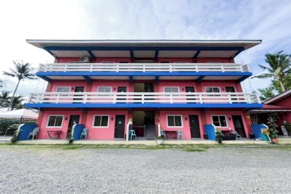 A two-story building with a pink and blue exterior, a balcony on the upper floor, and a gravel driveway. - Jamjen's Lodge (Baler)