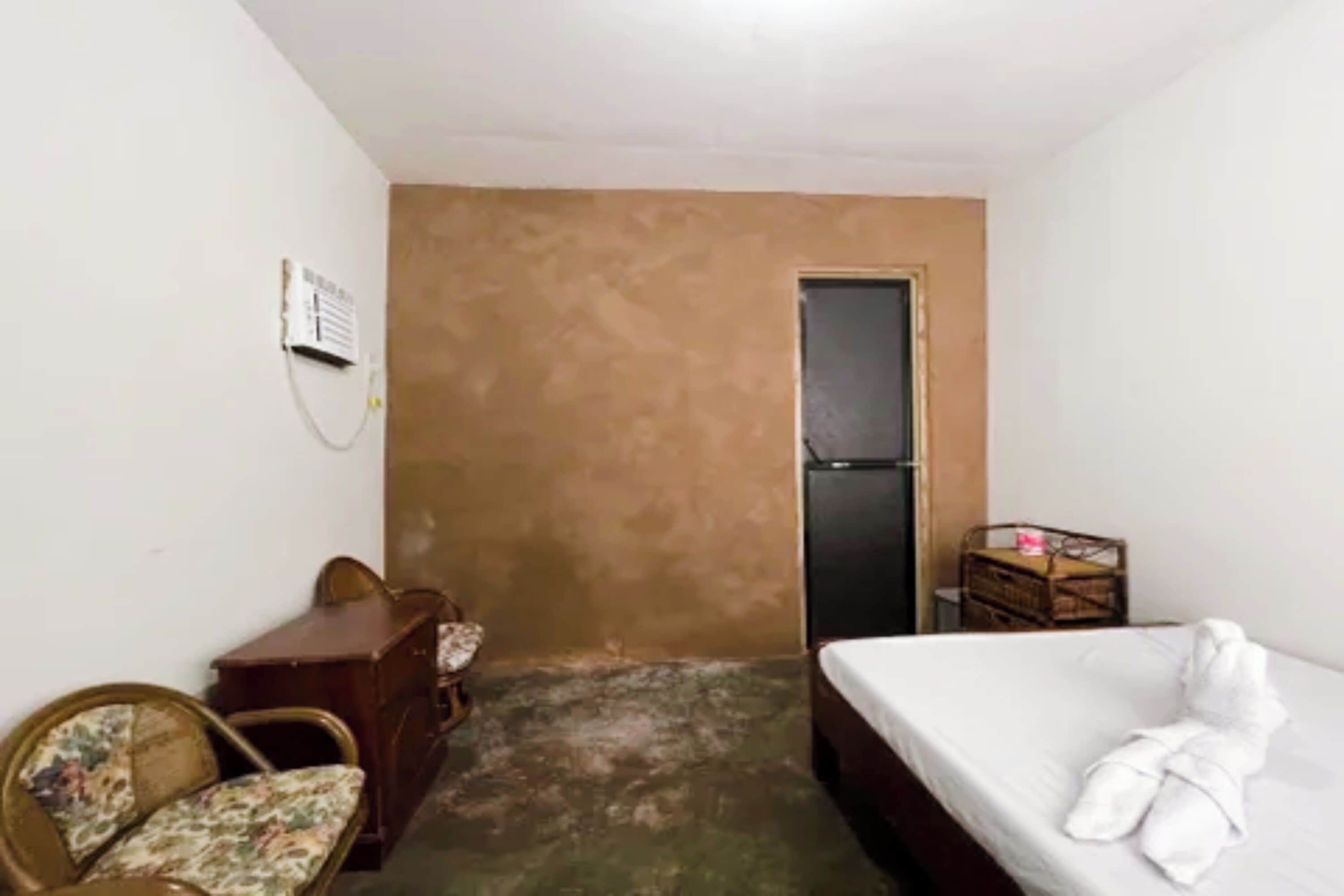 A small, sparsely furnished room with a bed, a wooden dresser, a chair, and a door.
