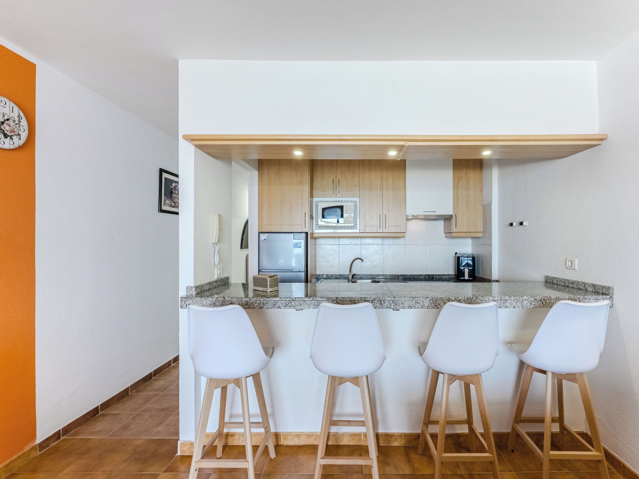 A compact kitchen with a central island, a refrigerator, and a microwave. There are four white bar stools around the island.