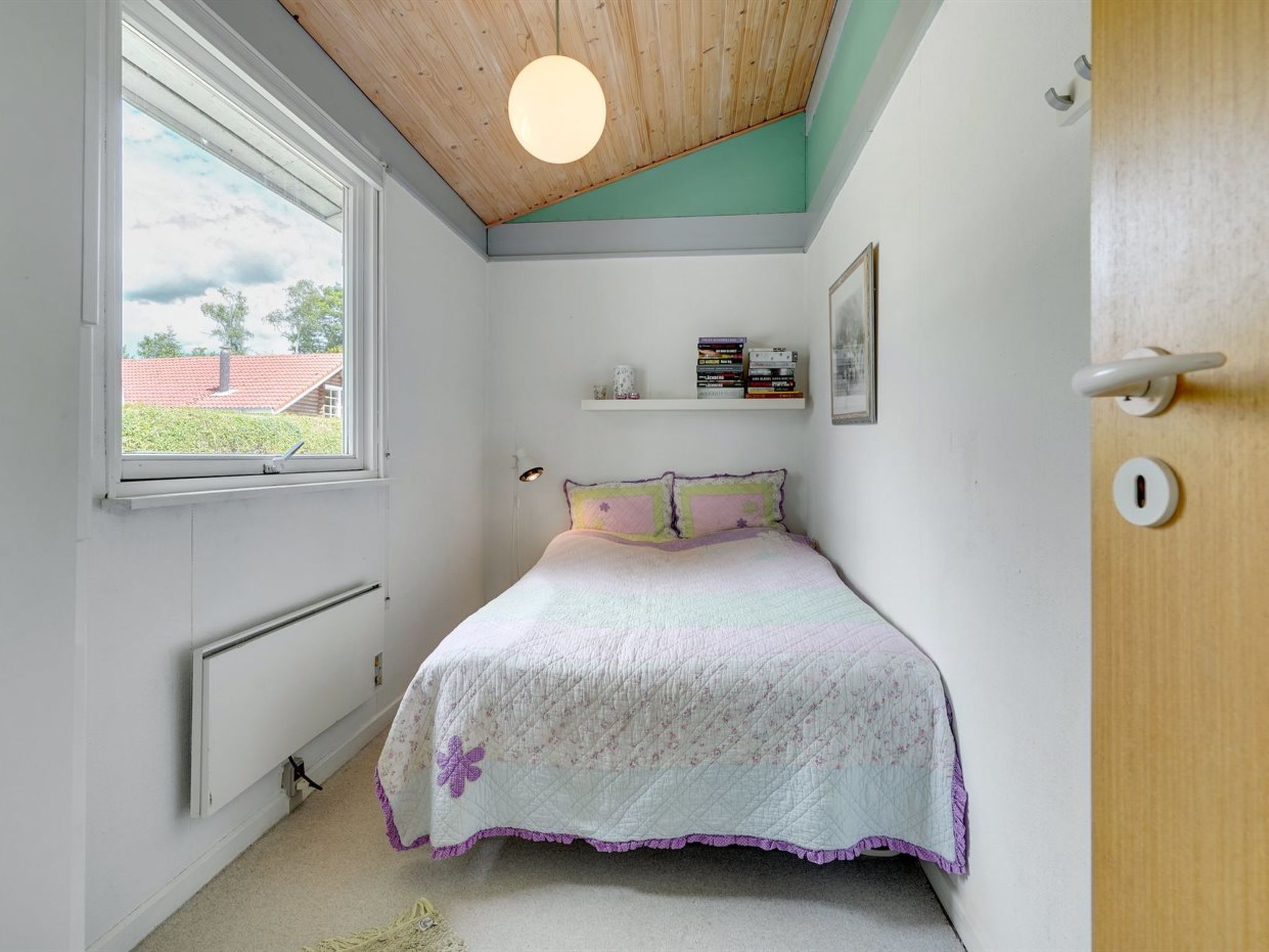 Una camera da letto con un letto, una finestra, un termosifone, una mensola con dei libri e una porta.