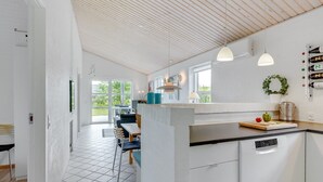 Moderni keittiö, jossa on valkoiset kaapit, musta työtaso ja laatoitettu lattia. Keittiössä on ruokailutila, jossa on pöytä ja tuolit, ja suurista ikkunoista avautuu näkymä ulos.