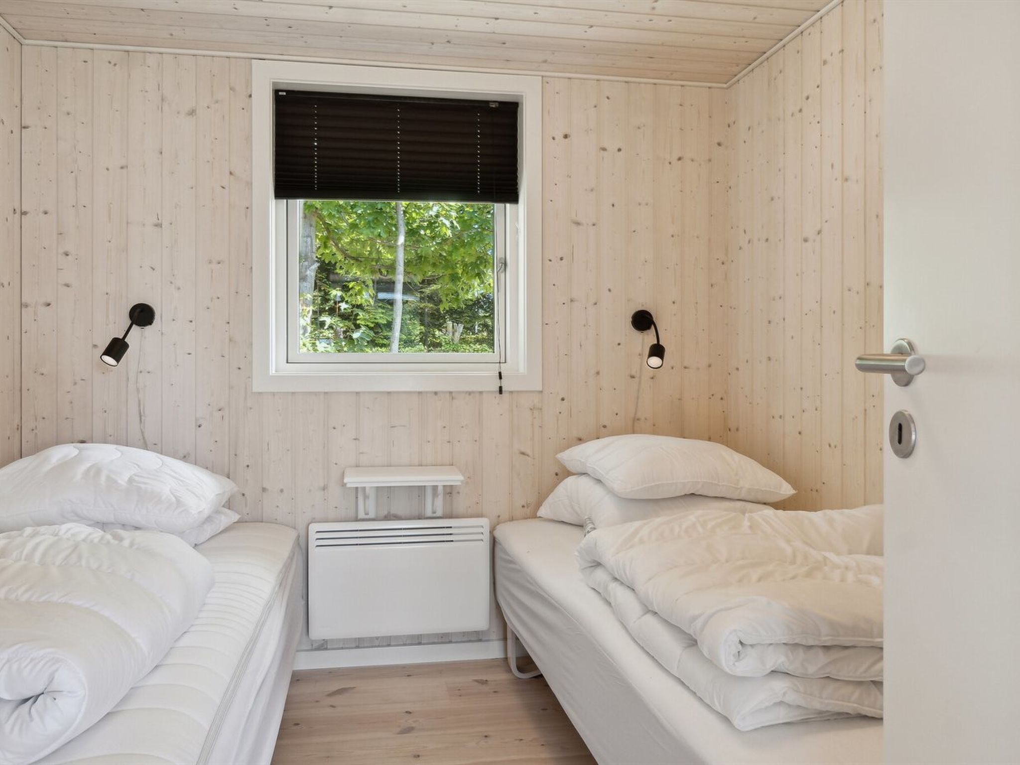 Una habitación con dos camas, una ventana con persianas, una mesita pequeña y luces en la pared.