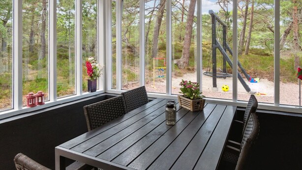 A covered patio with a wooden table and chairs, large windows offering a view of a playground and trees. - "Elsia" - 280m from the sea by Interhome (Nexø)
