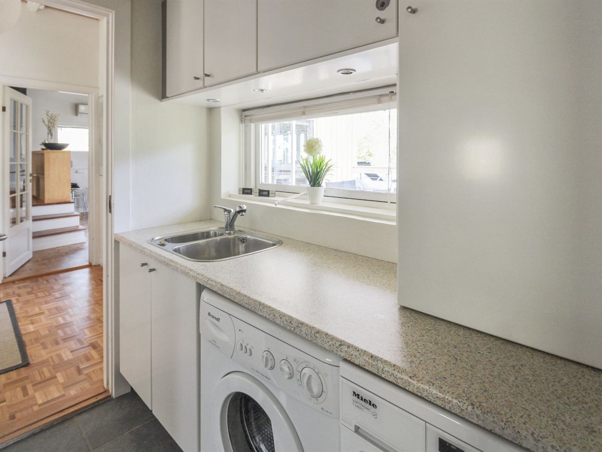 A compact kitchen with a built-in washing machine, a sink, and a window with a plant.