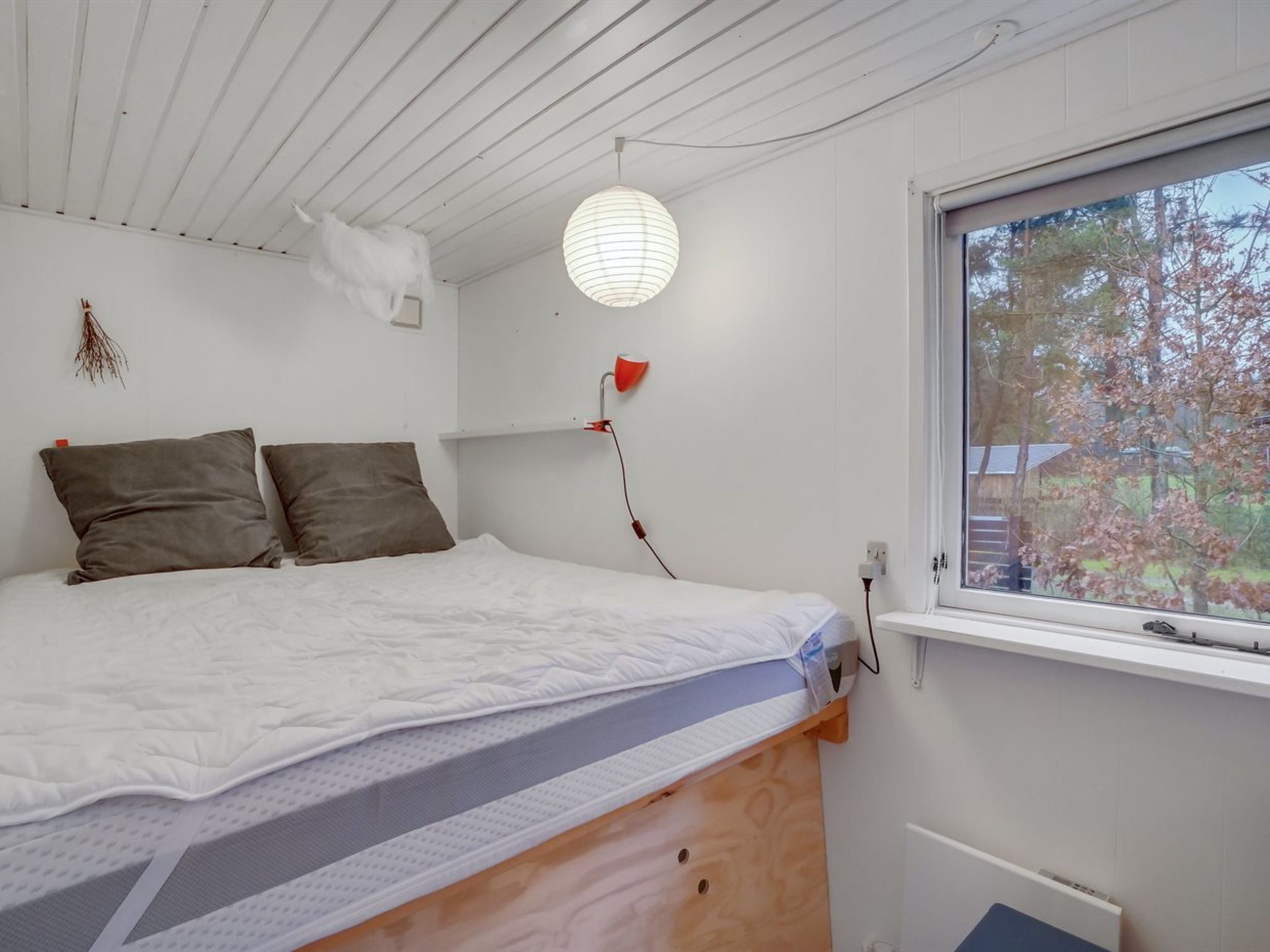 A bedroom with a bed, two pillows, a bedside lamp, a window, and a hanging light fixture.
