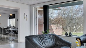 A modern living room with a leather sofa, a dining area with a table and chairs, and a large window with a view of trees and a brick wall. - "Markhild" - 300m to the fjord by Interhome (Hurup Thy)