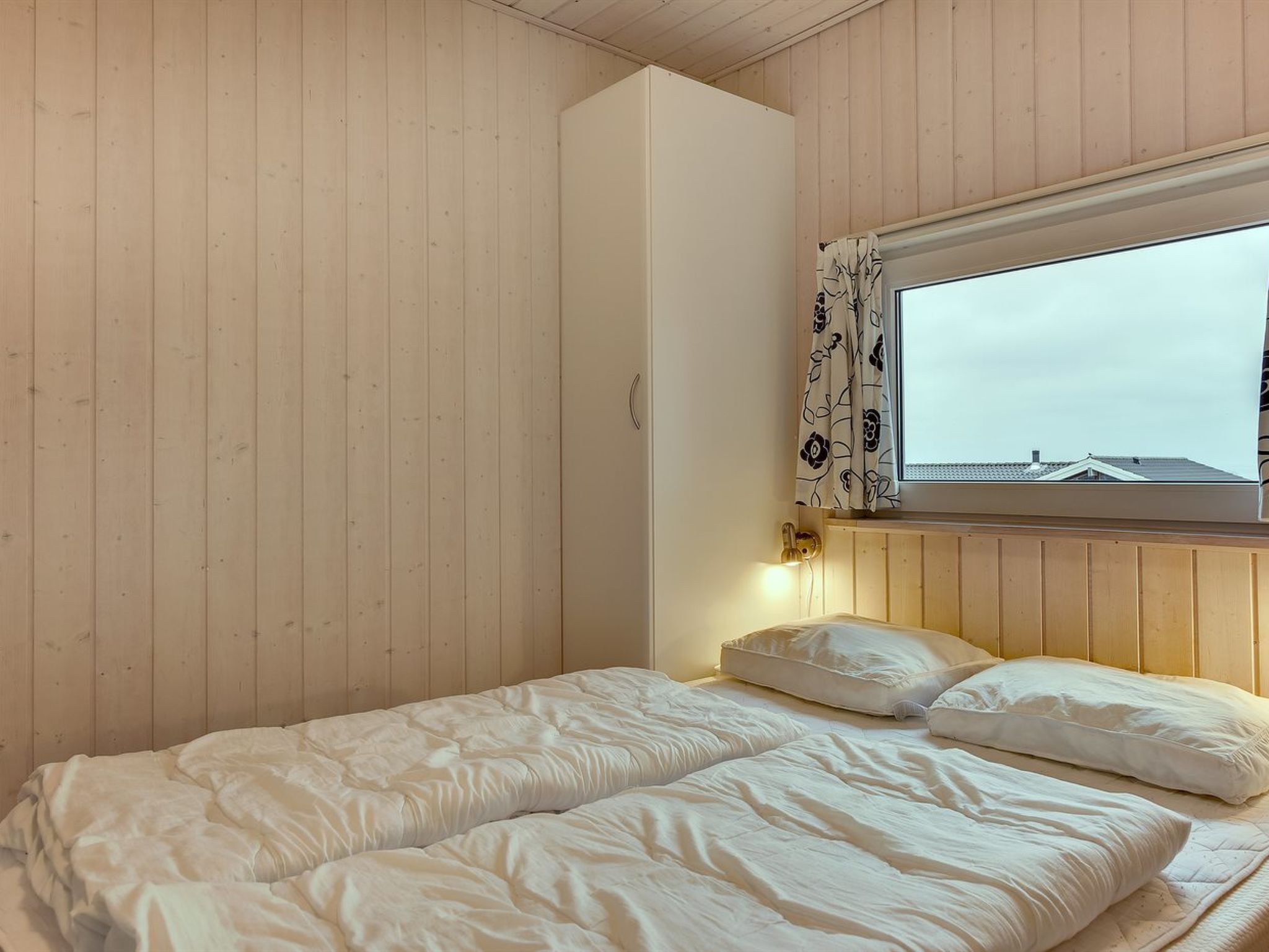 A bedroom with two beds, wooden walls, a window with floral curtains, and a wall-mounted light.