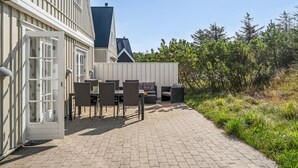 Een terras buiten met een eethoek, inclusief tafels en stoelen, grenzend aan een gebouw met een witte gevel en een donker dak.