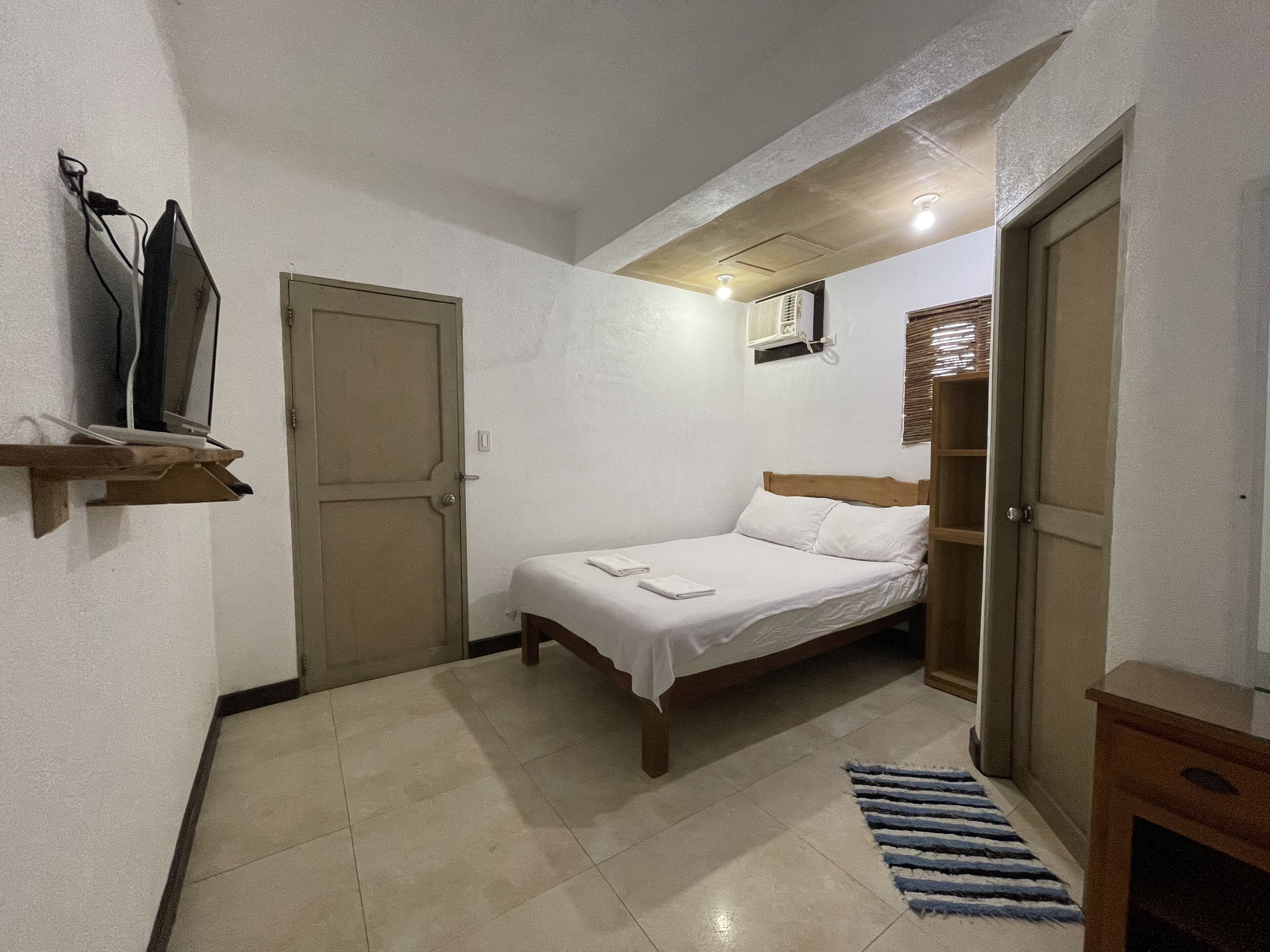 Un dormitorio pequeño y bien iluminado con una cama individual, cabecera de madera, un estante con un televisor y una puerta que da a otra habitación.
