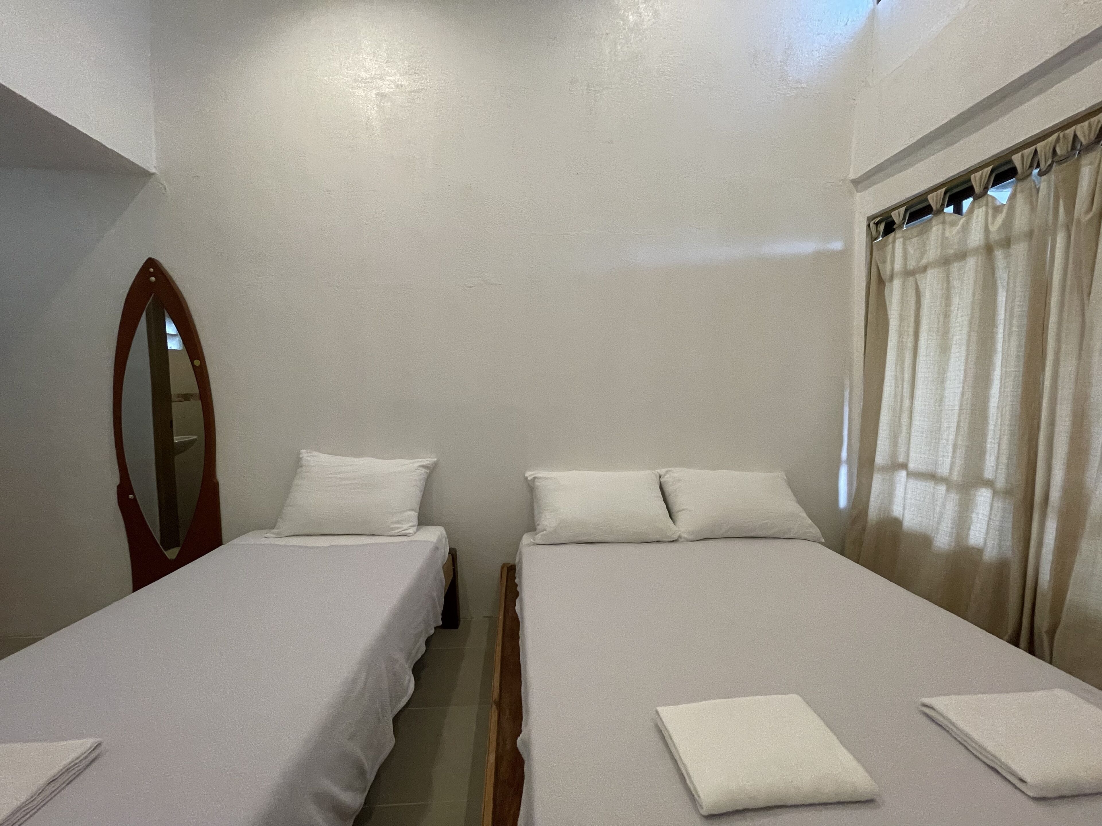 A room with two single beds, a wooden headboard, a mirror, and a window with curtains.