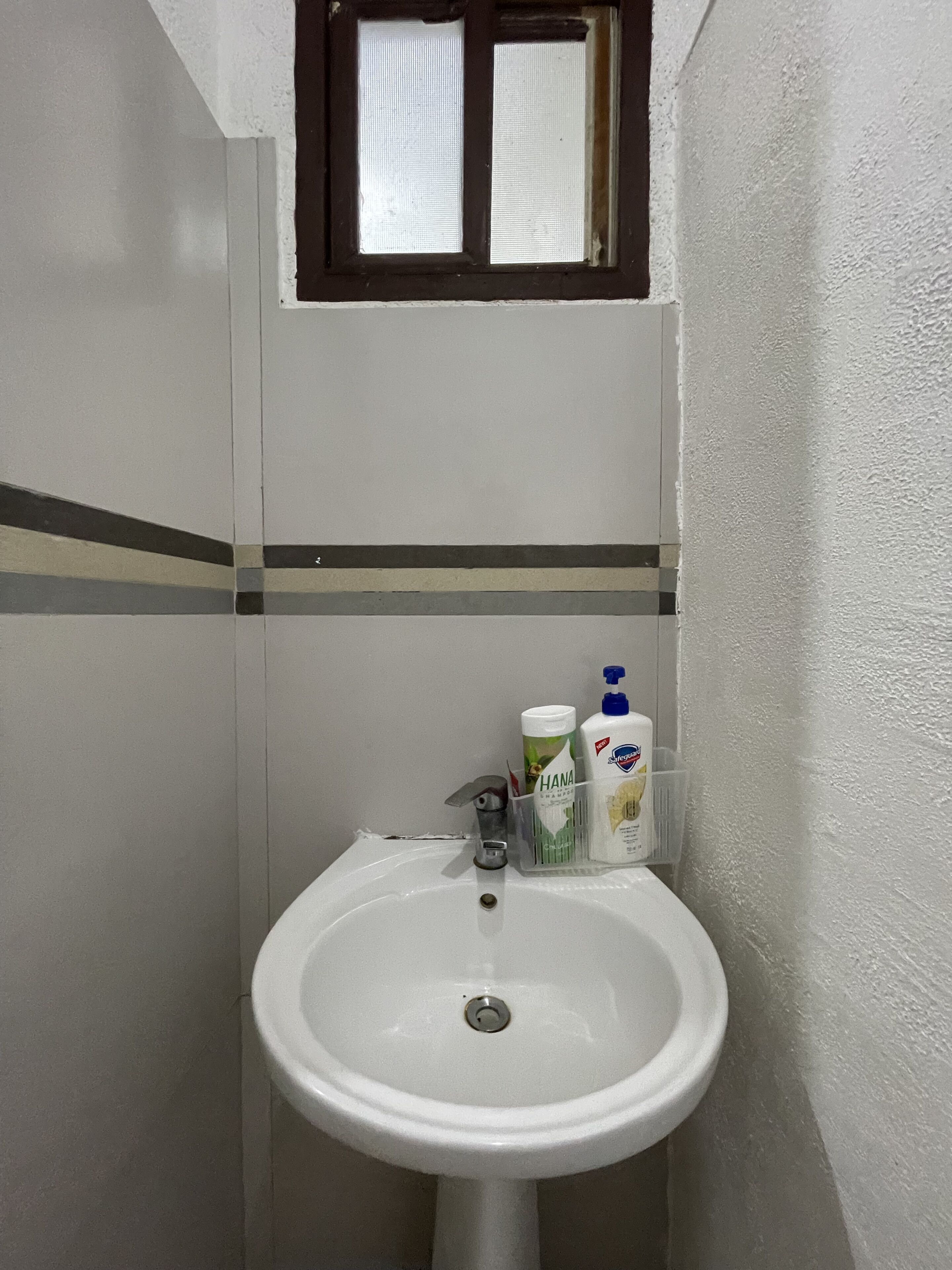 Un baño con un lavamanos blanco, una ventana encima y artículos de aseo sobre el lavamanos.