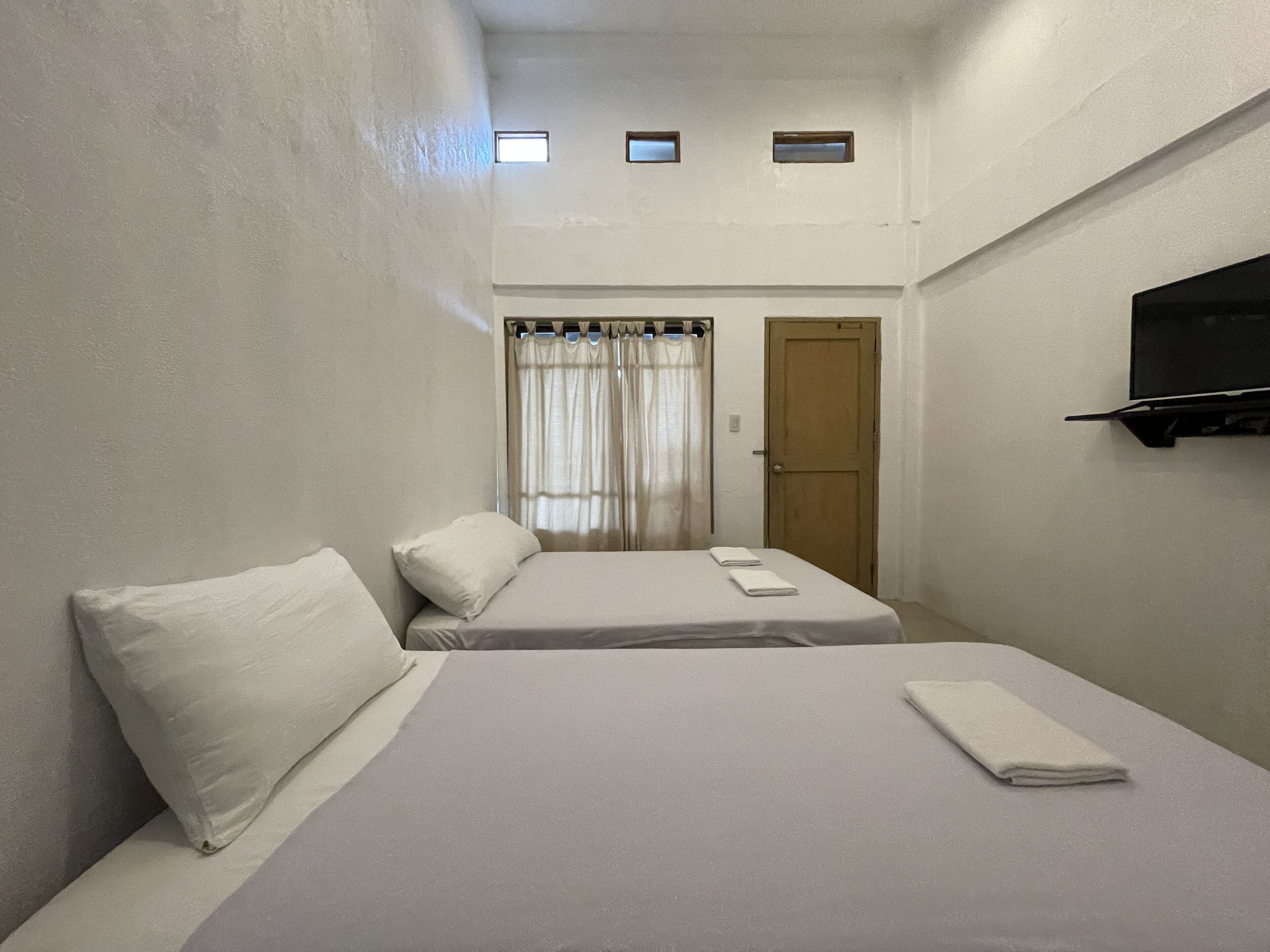 A small, clean room with two beds, a TV mounted on the wall, and a door leading to another room.
