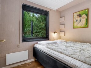 A bedroom with a bed, a window with a view of greenery, a bedside lamp, and a painting on the wall.