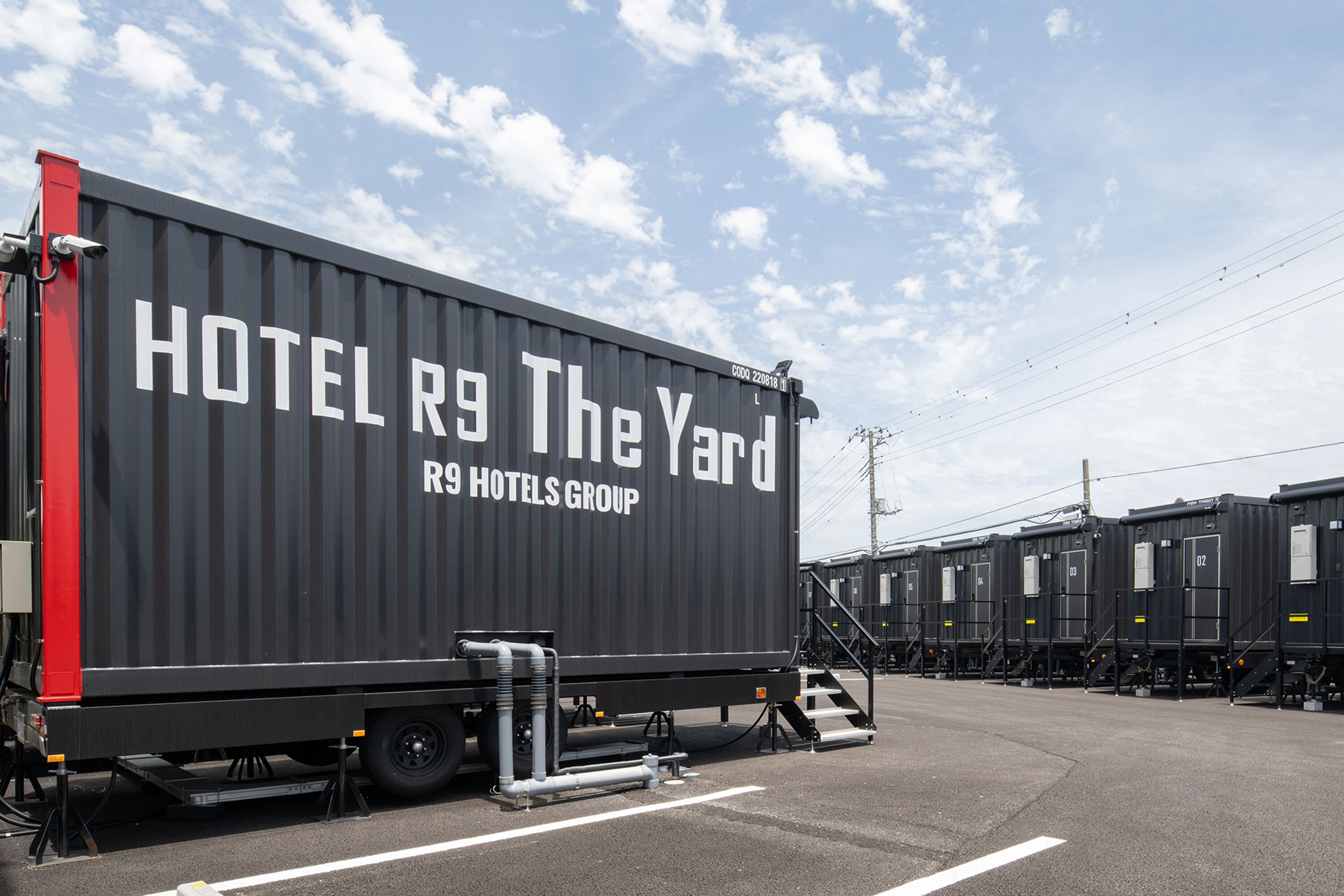 A large black container with "HOTEL R9 The Yard" and "RS HOTELS GROUP" written on it, parked in a lot with other similar containers.