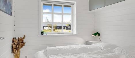 A bedroom with a large bed, a window with a view of the outside, a green desk lamp, and a vase with dried plants.