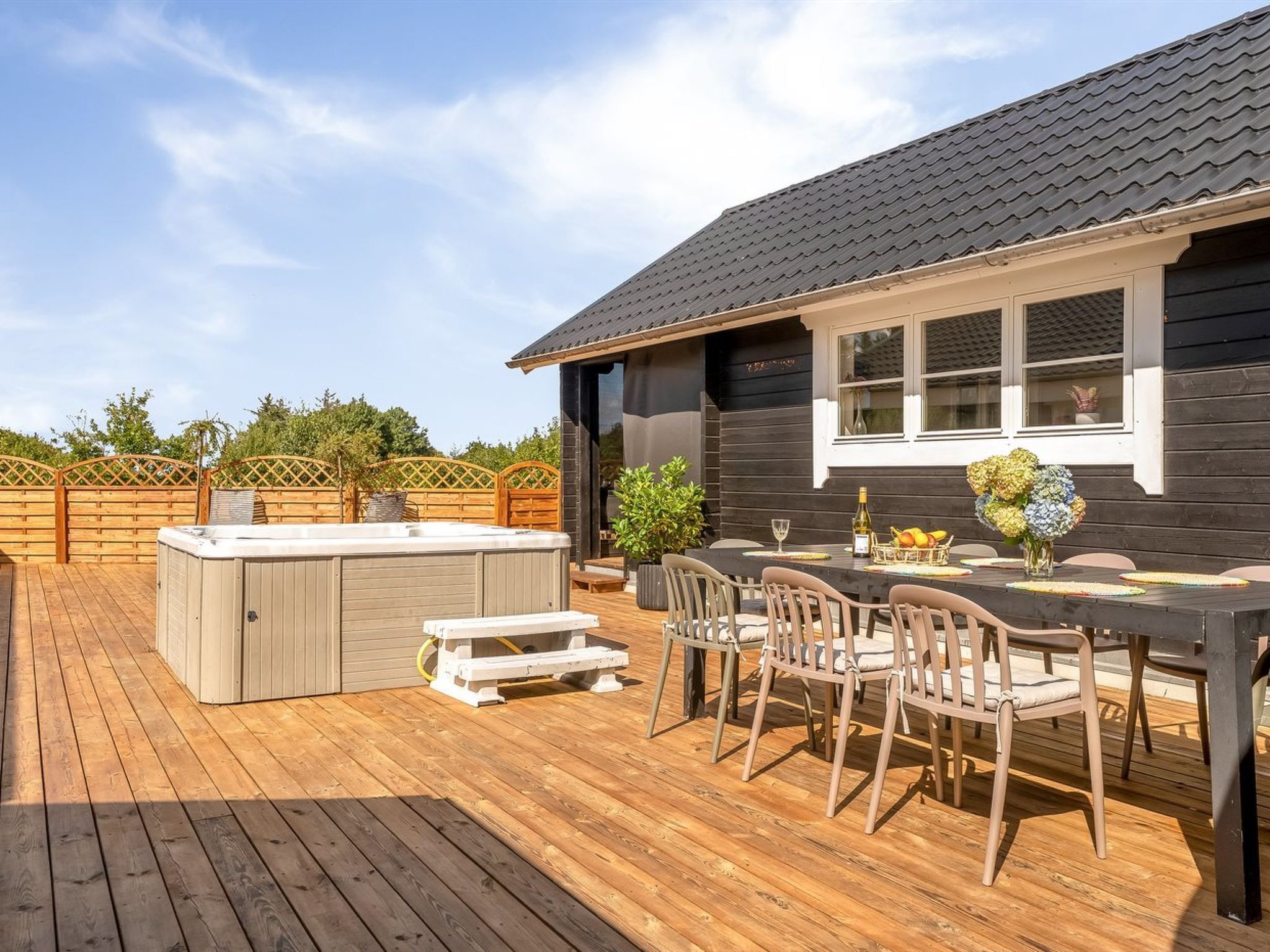A wooden deck with a hot tub, outdoor seating, and a house with a dark roof and windows.