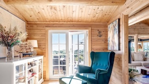 Ein gemütliches Blockhaus-Interieur mit Holzwänden, einem großen Fenster, einem türkisfarbenen Sessel und einem Bücherregal.
