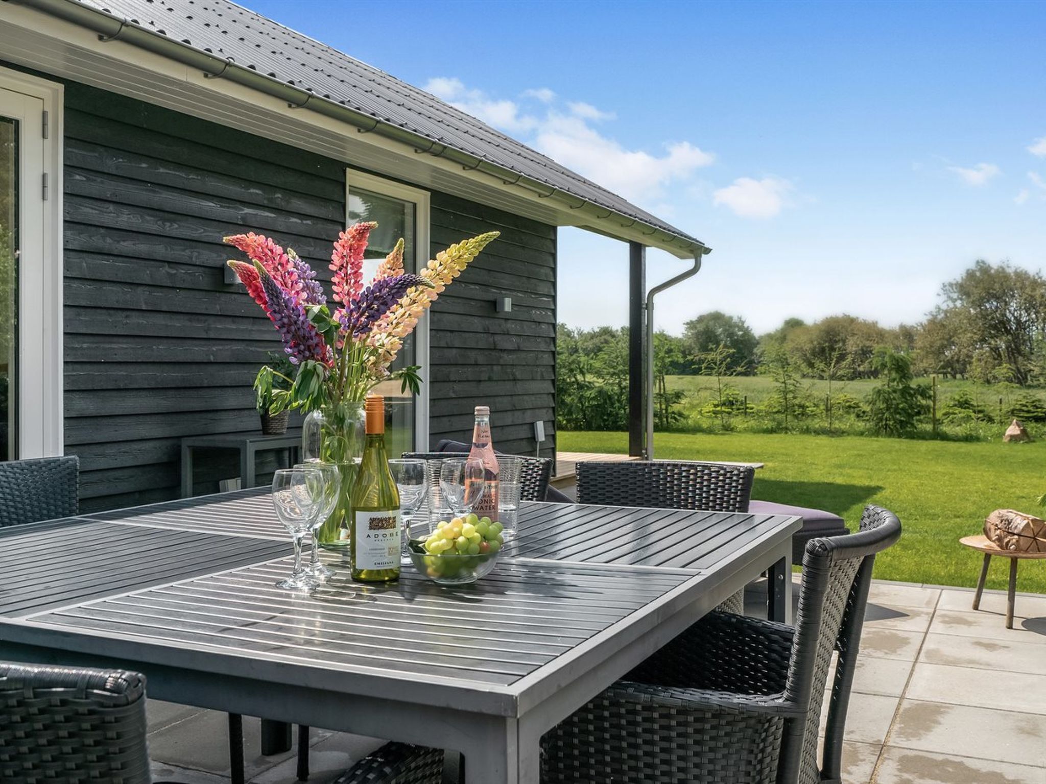 Une terrasse extérieure avec une table dressée pour le dîner, agrémentée d’une bouteille de vin, de verres et d’un bol de raisins.