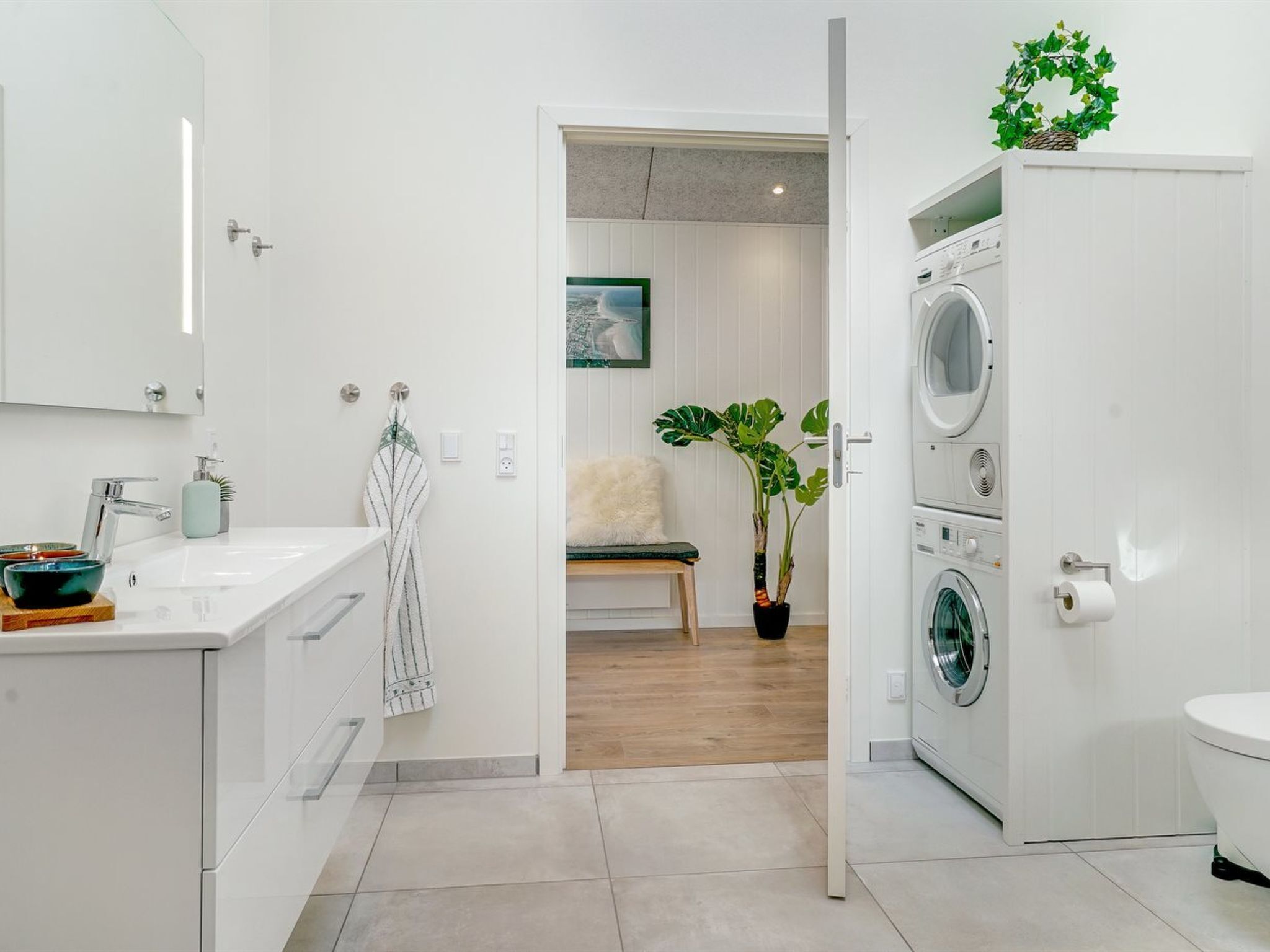 Une salle de bain moderne avec un lavabo blanc, un miroir et un lave-linge. On aperçoit, au fond, une plante en pot et une chaise.