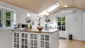 A modern kitchen with white cabinetry, a wooden countertop, and a built-in island with glass doors. There are two pendant lights, a window with a view of greenery, and a clock on the wall. - "Engelbertine" - 210m to the fjord by Interhome (Struer)