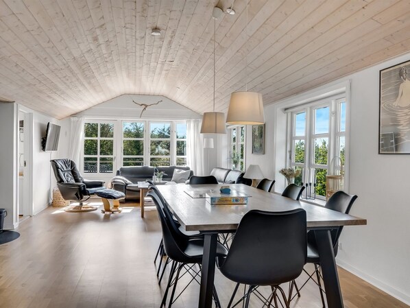 A spacious dining area with a long table and multiple chairs, a sofa, and a television mounted on the wall.