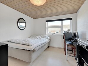 Une chambre à coucher avec un lit, un bureau avec un ordinateur, une chaise, un miroir et une fenêtre donnant sur des immeubles.