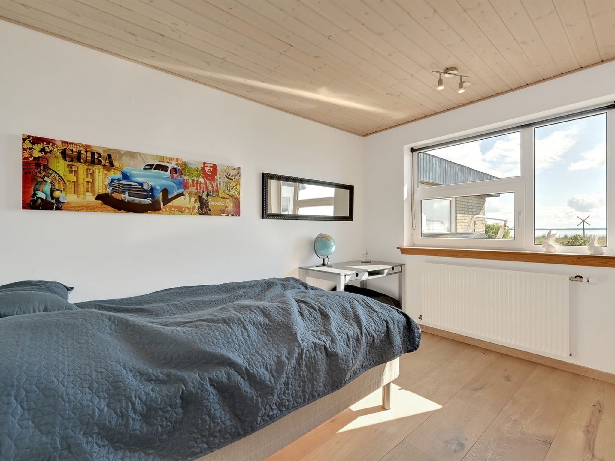 A bedroom with a bed, a desk, a globe, and a window with a view of the sky and wind turbine.