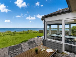 Een terras met tafel en stoelen, uitzicht op zee, en een gebouw met grote ramen.