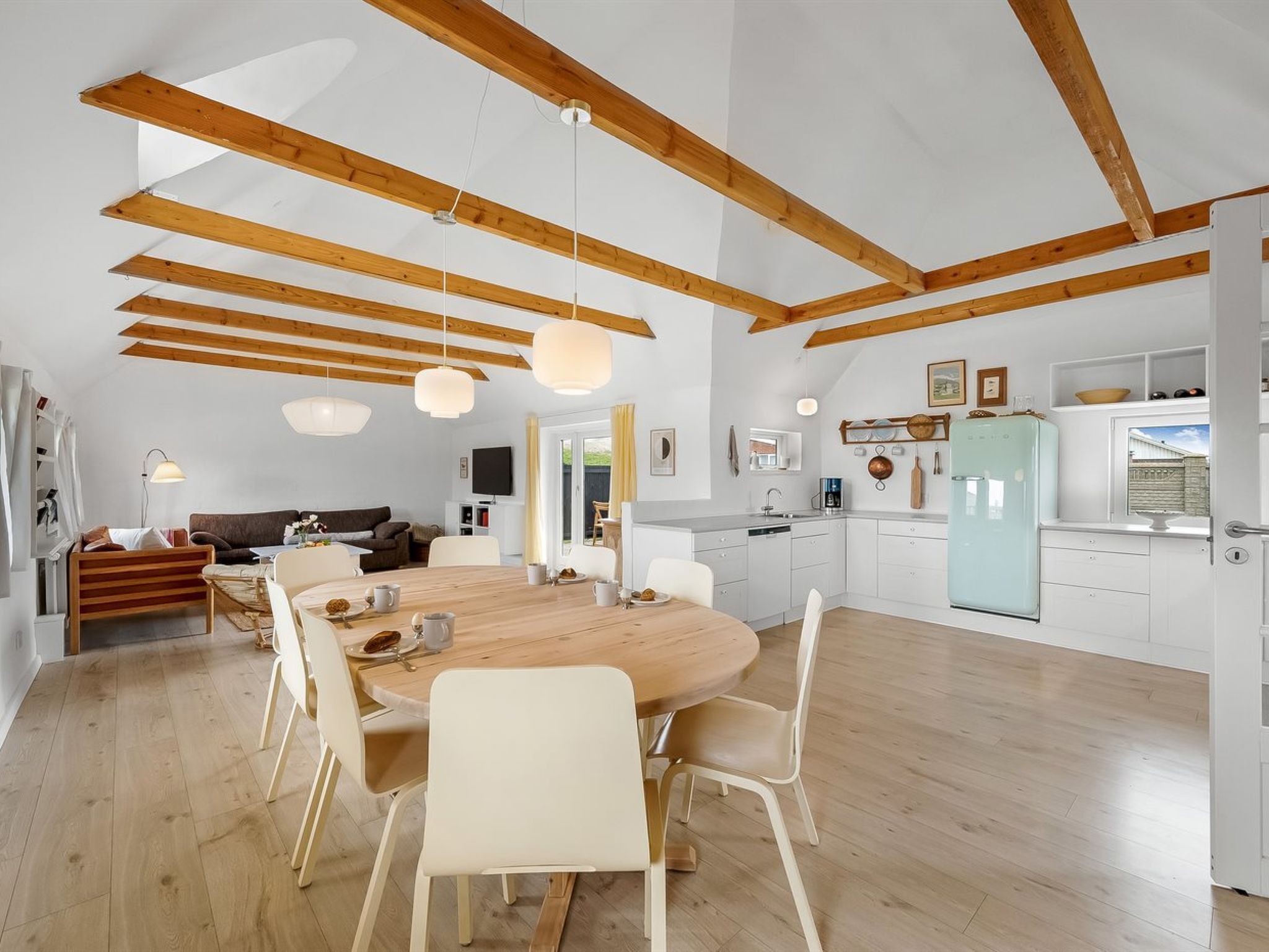 A spacious kitchen with wooden beams, a dining area with a round table and chairs, and a modern kitchen setup including a fridge and cabinets.