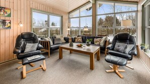 A cozy room with wooden walls and large windows, featuring a sofa set, a coffee table, and leather chairs.