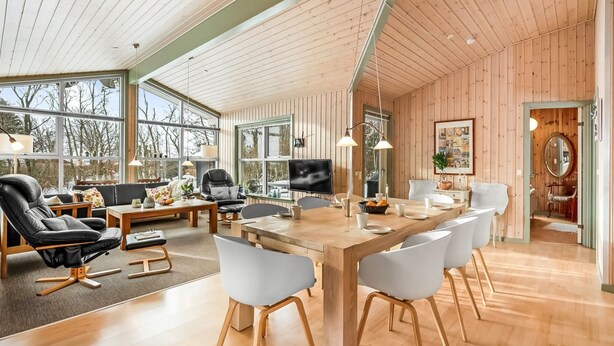 A spacious living room with a wooden ceiling, large windows, a dining area with a long table and chairs, and a cozy seating area with a sofa and chairs.