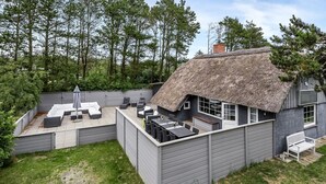 Une maison moderne avec un toit de chaume, un revêtement extérieur gris et un patio aménagé avec du mobilier d’extérieur.