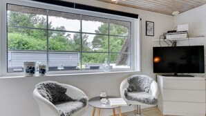 Une chambre moderne dotée d’une grande fenêtre, de deux chaises blanches recouvertes de coussins en fausse fourrure grise, d’une petite table, d’un téléviseur et d’un meuble blanc.