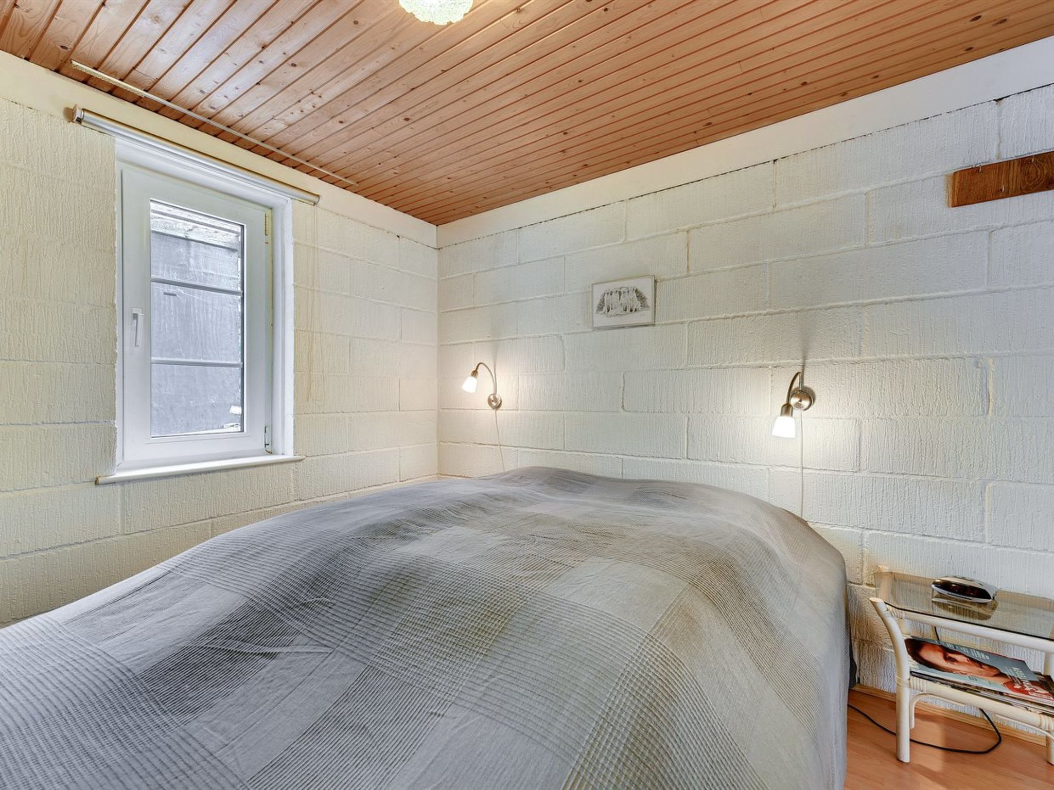 Une chambre avec un plafond en bois, un lit recouvert d’une housse grise, une fenêtre et deux lampes fixées au mur.