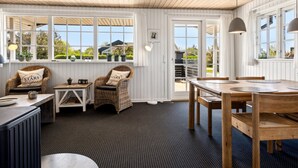 A room with a wooden ceiling, white walls, a dining table, wicker chairs, a small table with a lamp, and large windows with a view of greenery outside. - "Ekatarina" - 125m from the sea by Interhome (Juelsminde)