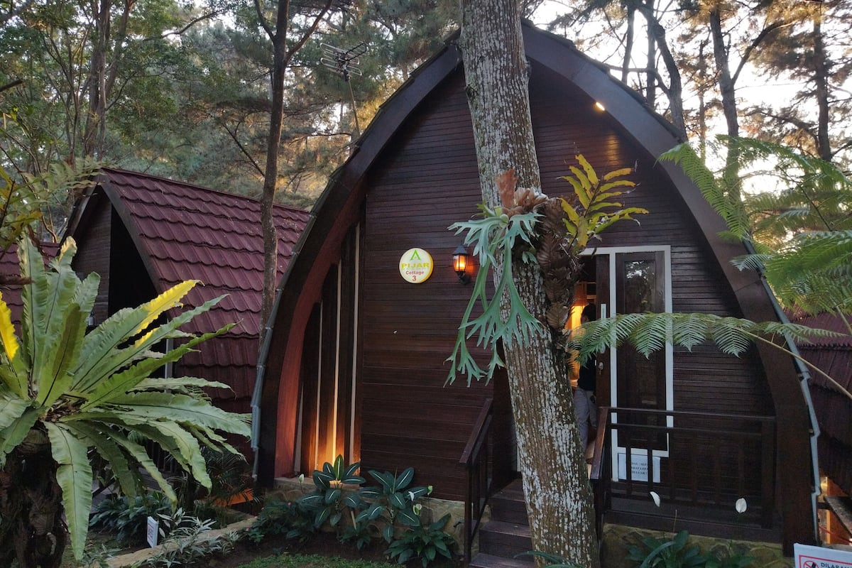 A wooden cottage with a red roof, surrounded by lush greenery and a sign indicating it is a 'Plaza Cottage 3'.