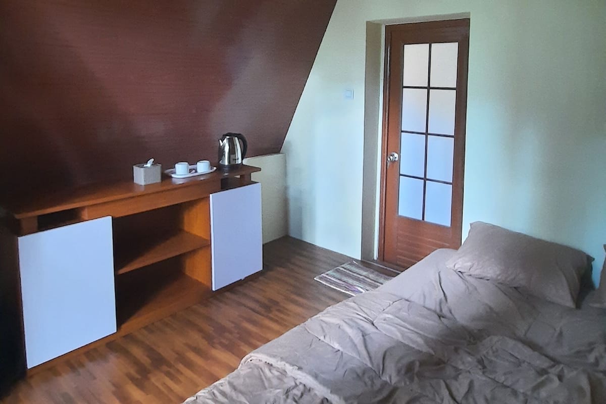 A bedroom with a sloped ceiling, a bed, a desk with a chair, a small table with a kettle, and a door with a window.