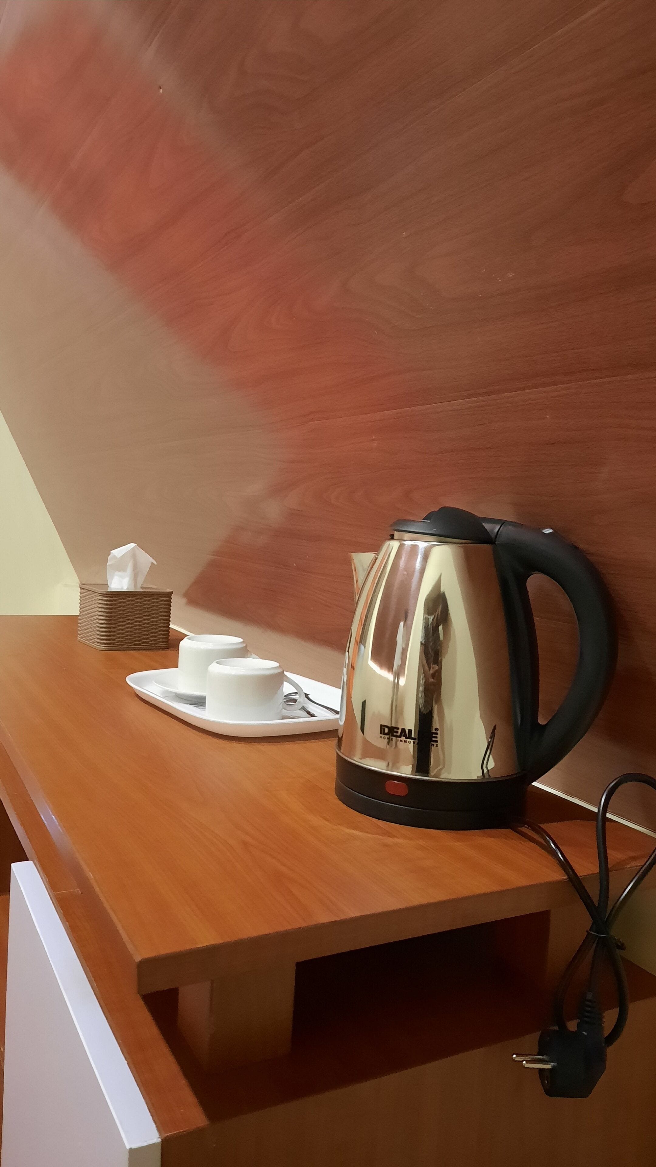 Una scrivania in legno con un bollitore in acciaio inox, un scatolo di fazzoletti e due tazze bianche.