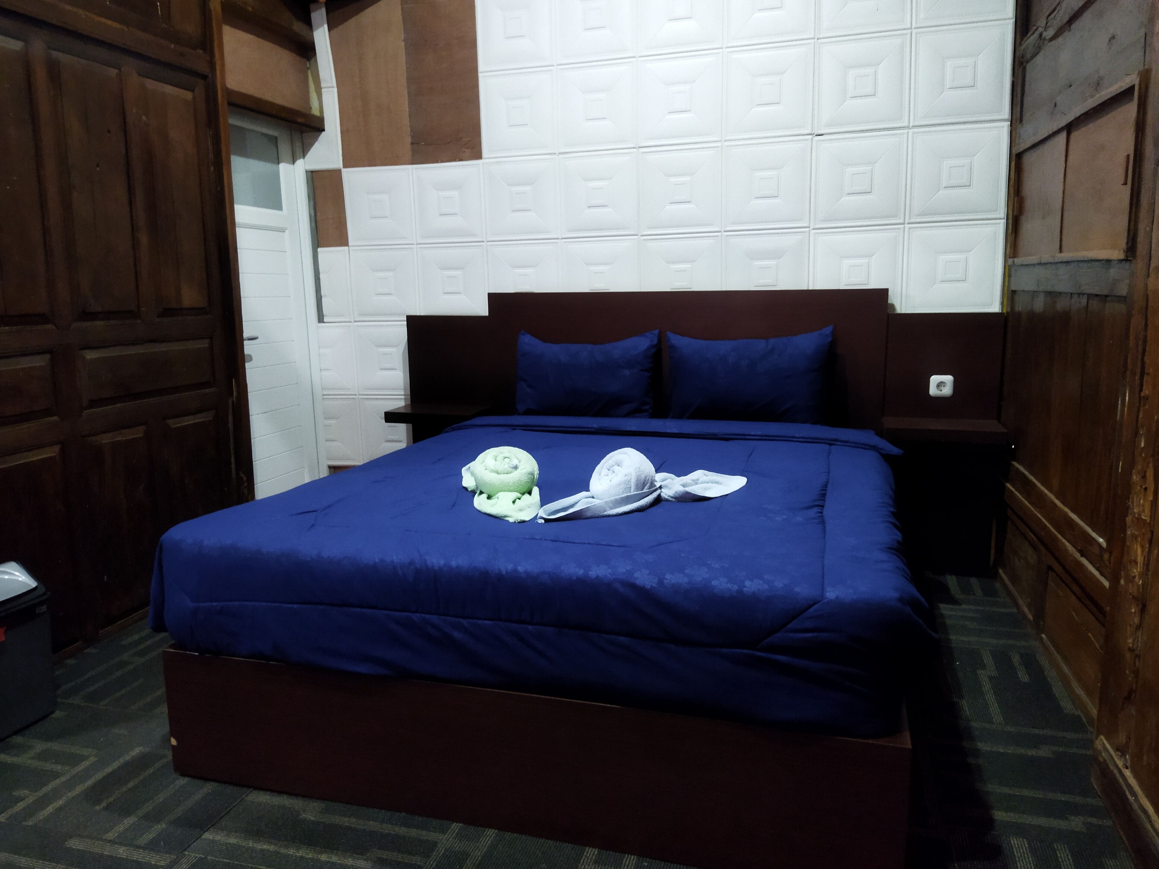 A hotel room with a large bed, a wooden headboard, and a white tiled wall.