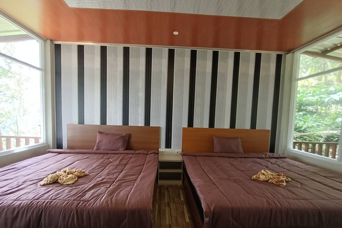 A room with two beds, each with a wooden headboard and a purple bedspread. There is a window on the left side, and a curtain with vertical stripes is visible behind the beds.