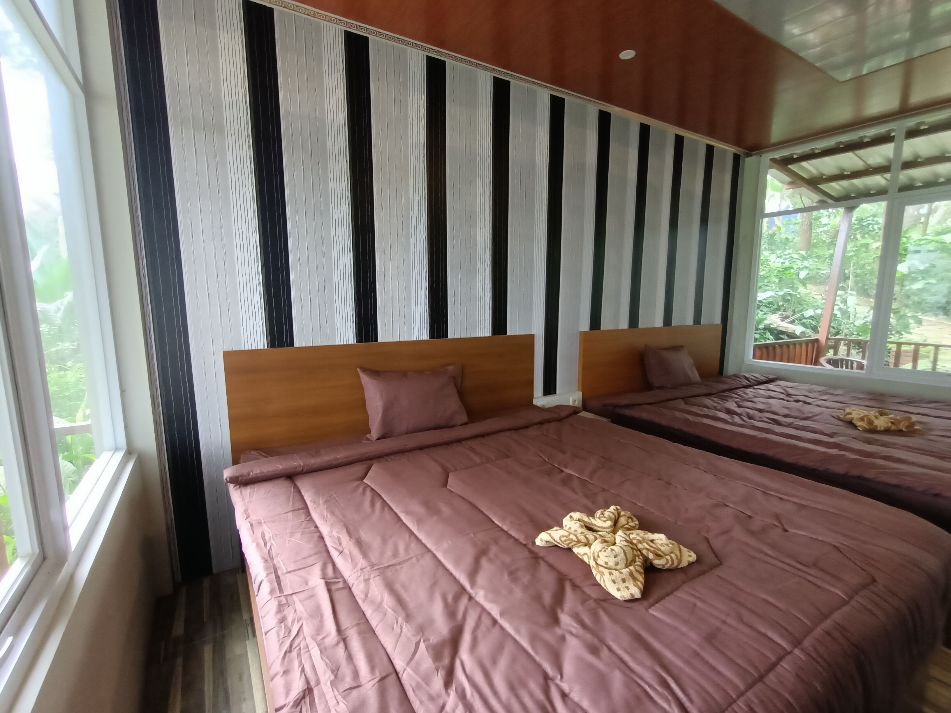 A room with two beds, each with a wooden headboard and a pink bedspread. There is a window on the left side, and a view of greenery outside.