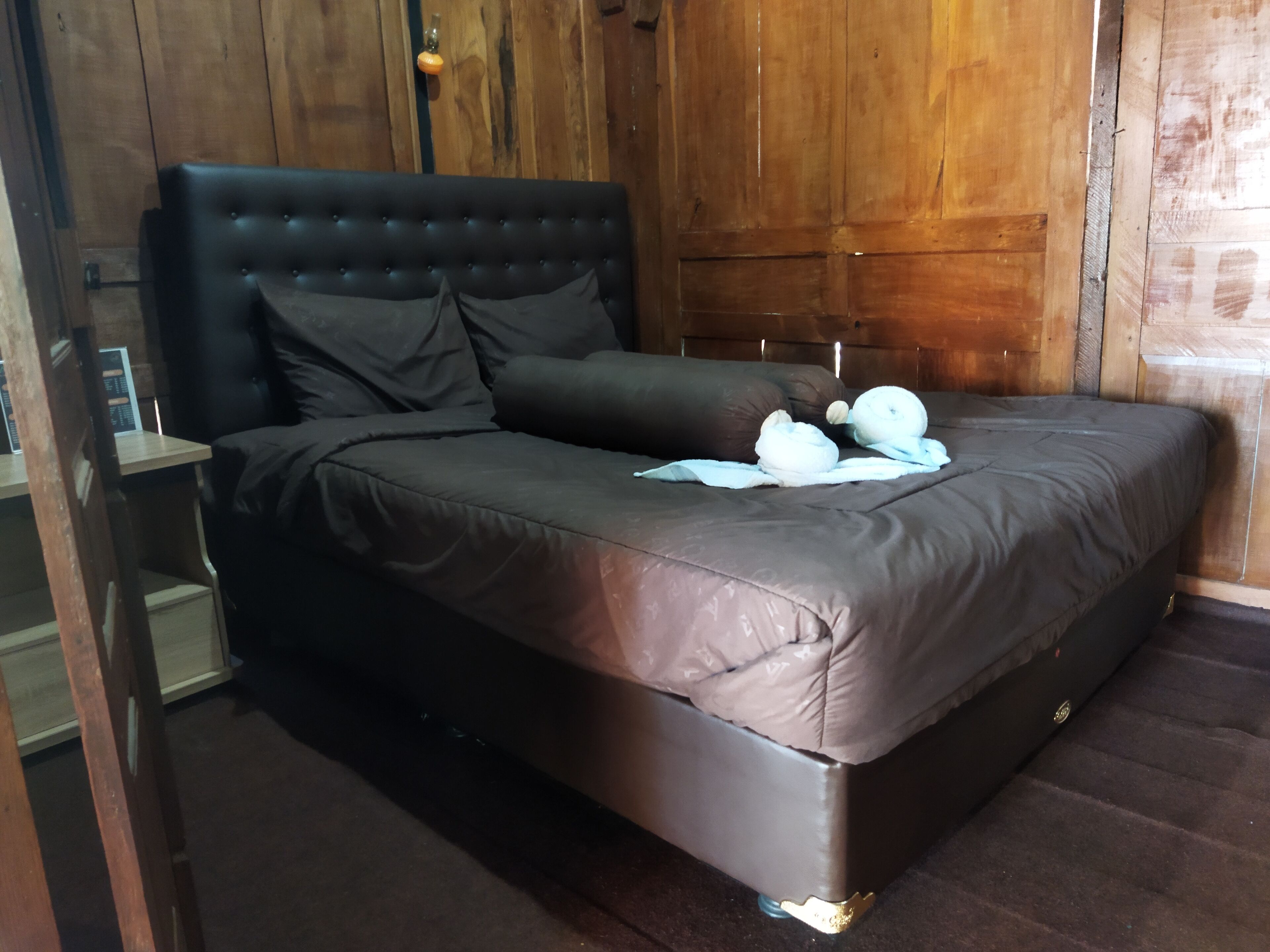 Una cama con cabecera oscura y almohadas, una mesita de noche pequeña y una pared de madera.