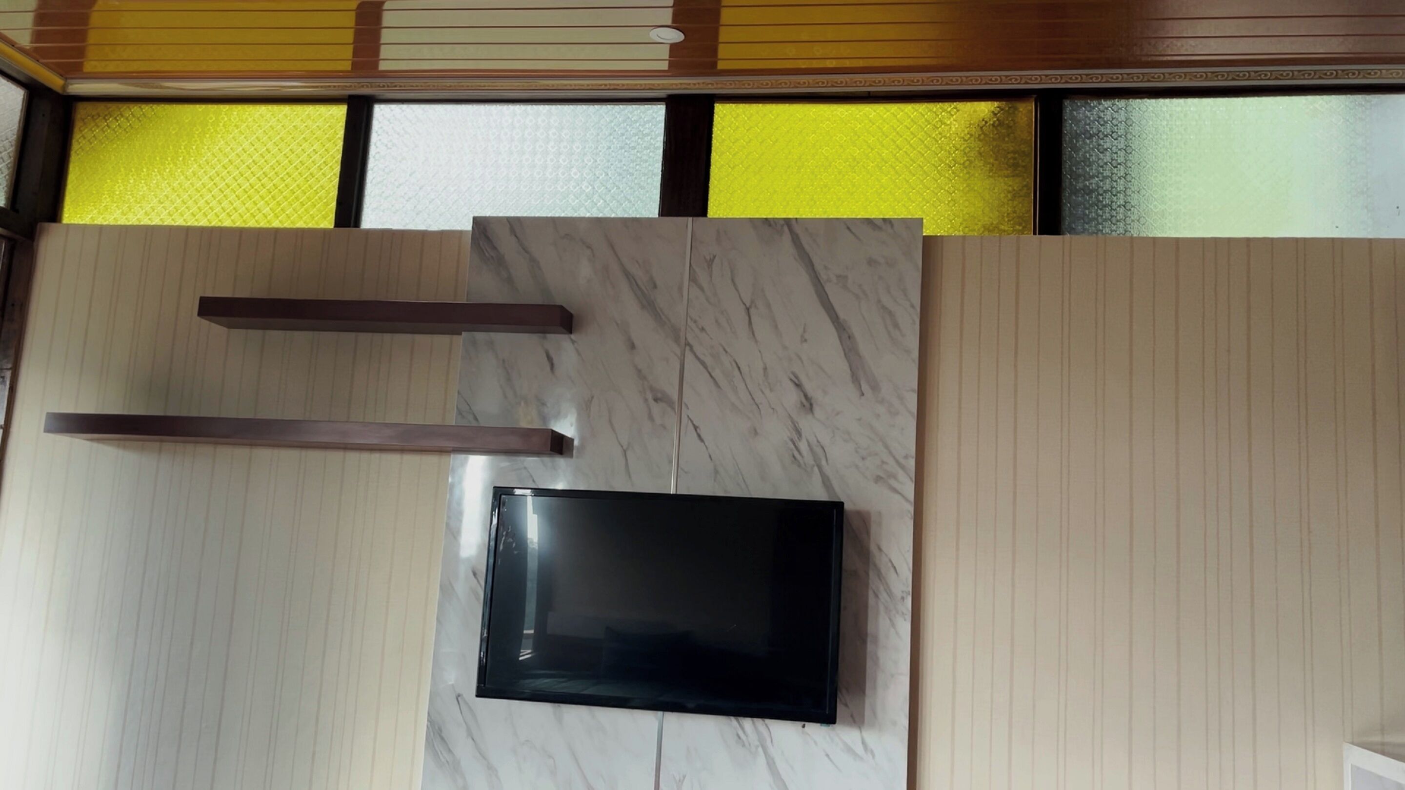 Quarto com uma grande TV de tela plana sobre uma superfície de mármore, lambris de madeira e janela com padrão amarelo.