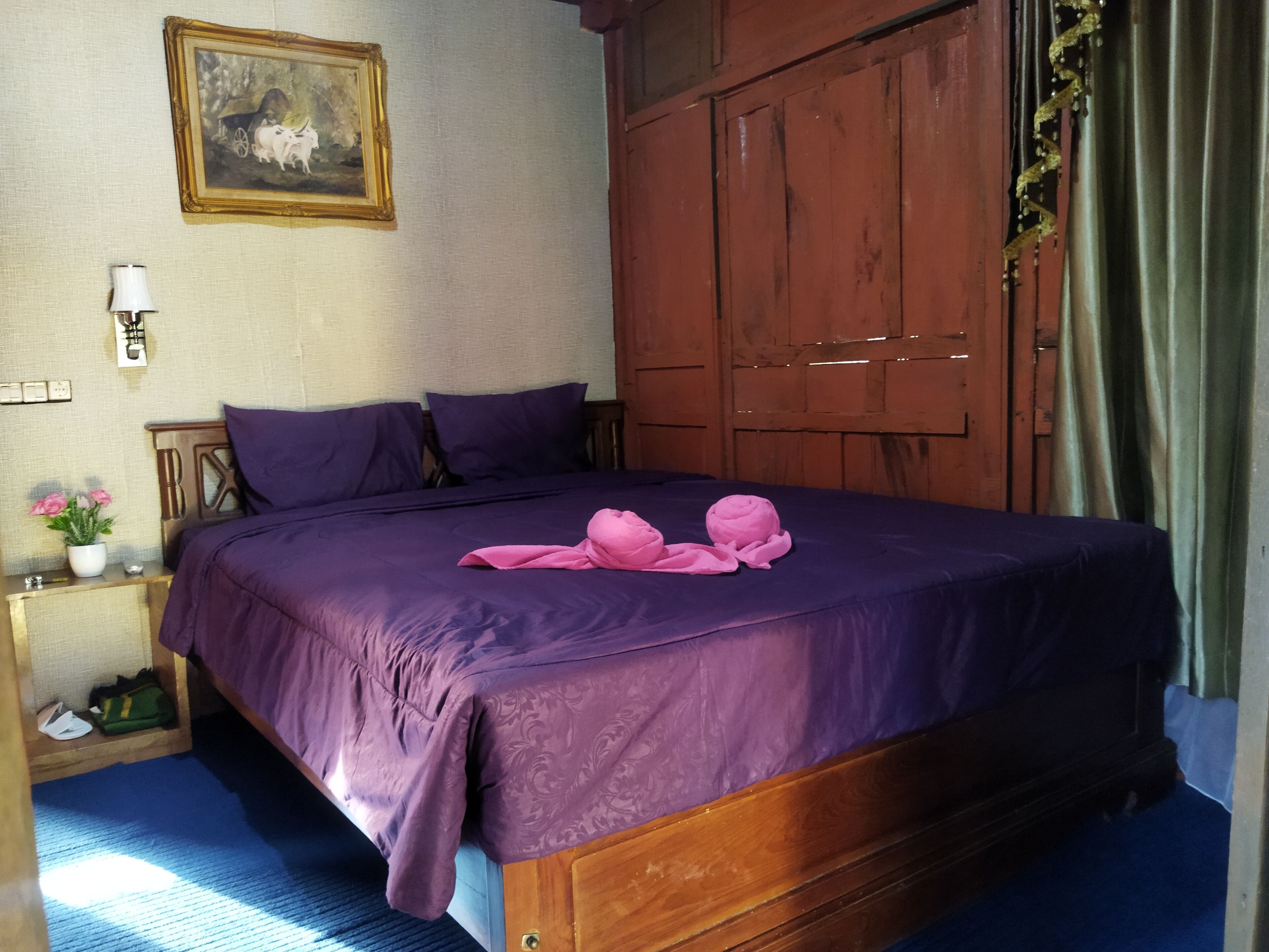 A bed with purple bedding, a wooden wardrobe, a nightstand with a lamp, and a framed picture on the wall.